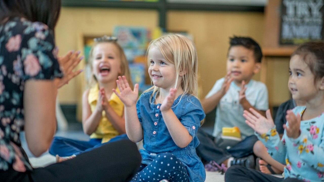 Children sitting on the floor in a classroom, smiling and clapping along with an adult.
