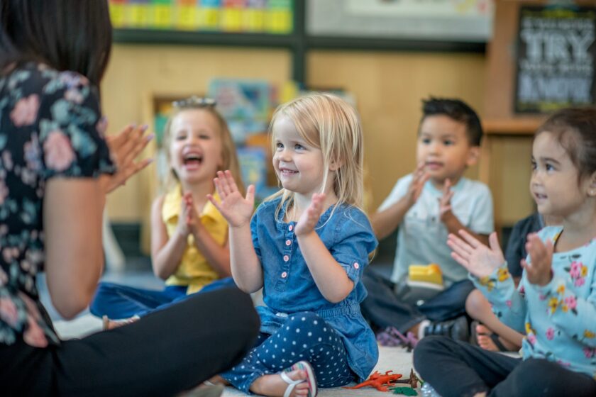 Children sitting on the floor in a classroom, smiling and clapping along with an adult.