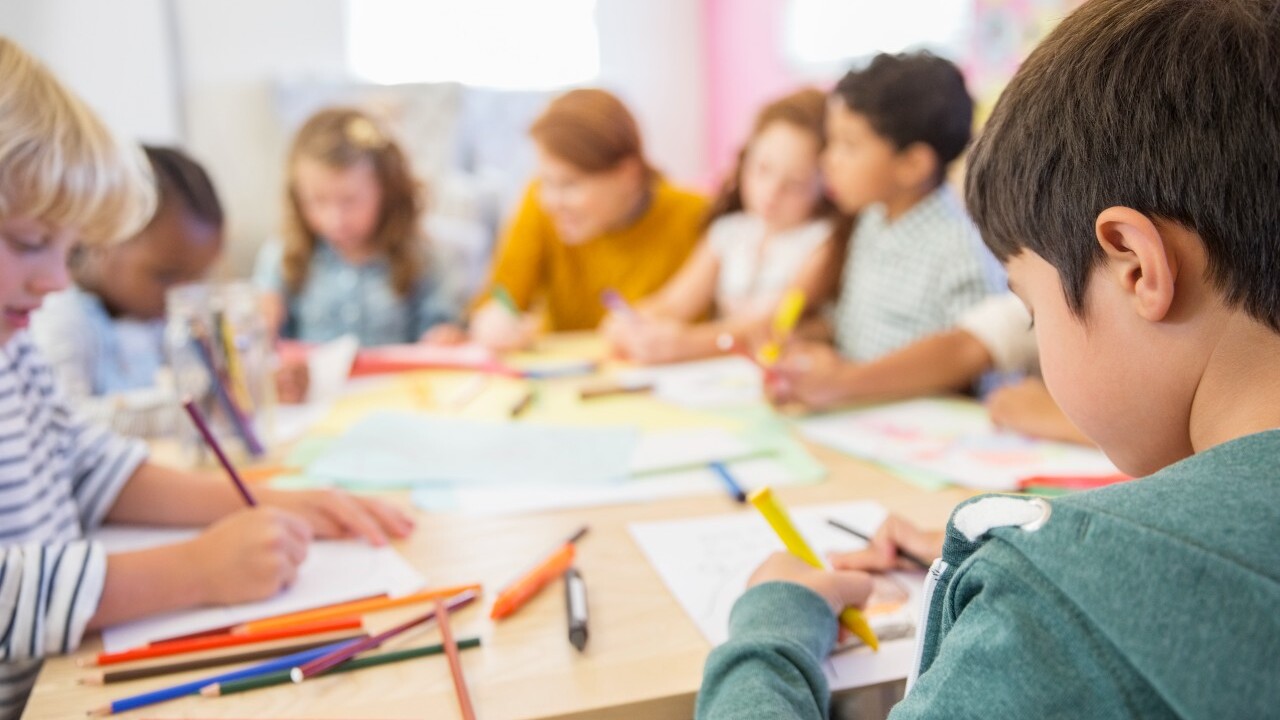 Children sitting at a table, focused on drawing with colored pencils and markers on paper. An adult is assisting in the background.